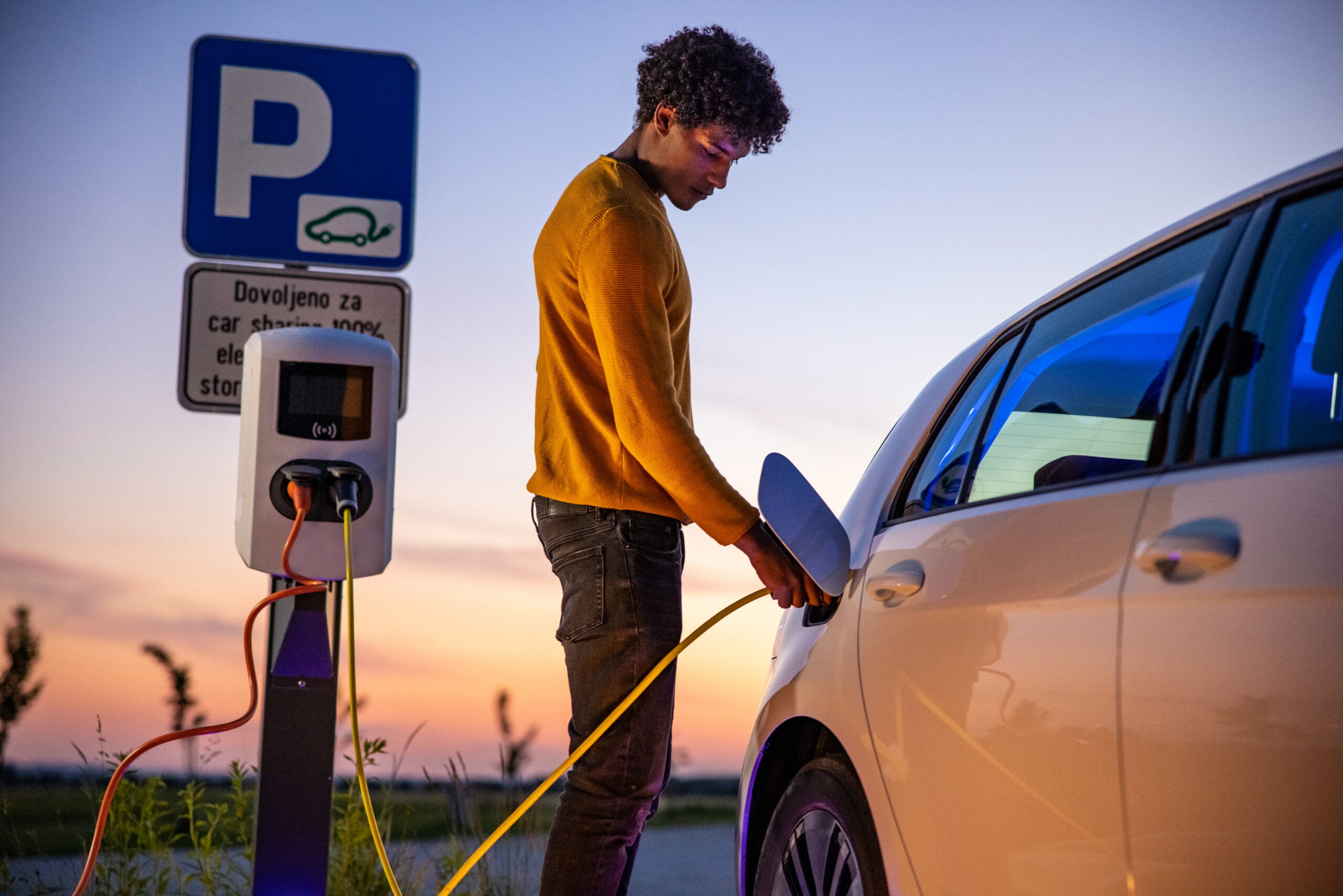 Young black man with curly hair charging his electronic car at a charging station during blue hour