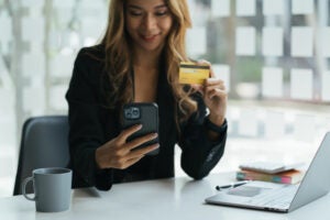 A young asian business woman its at her desk with her computer open while using her smart phone and holding up her credit card.