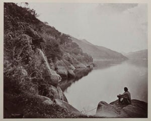 A black and white photo of a man sitting on rocks gazing down at the river