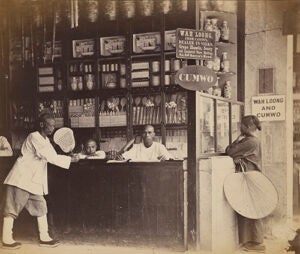 An old black and white photograph of a shop counter with sellers and buyers gathered around it.