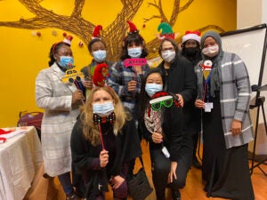 A diverse group of women wearing Christmas themed accessories and masks smile at the camera.