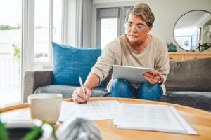 A senior woman with short hair holds and iPad while leaning over a coffee table tot make notes on papers spread across it.