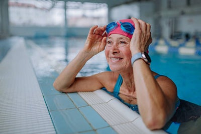Elderly woman in a swimming pool lifts her goggles and smiles into the distance.