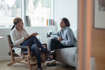 A woman sitting on a couch speaks to her therapist, who listens holding a clipboard ad pen.