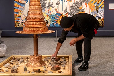 In this photograph, a man bends over to pour debris at the base of a mandala-shaped sculpture. 