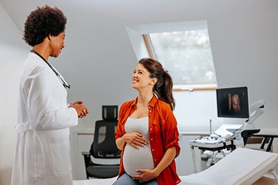 A pregnant woman in a red open-collared shirt smiles at a doctor in a lab coat. 