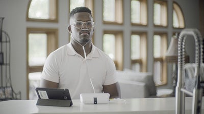 A black man in a white polo shirt stares into the distance, utilizing a device that helps stabilize his breathing by connecting to a nasal cannula.