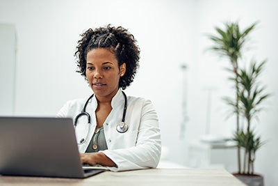 A female provider wearing a white coat and a stethoscope around her neck types on a gray laptop. 