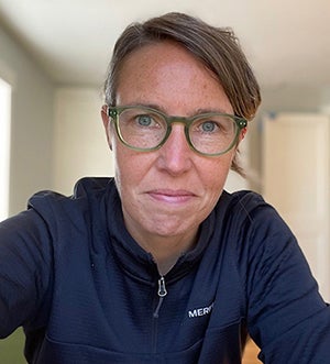 Selfie of a freckled woman with light blue eyes wearing green glasses and a navy blue athletic zip-up sweater while smiling slightly at the camera.