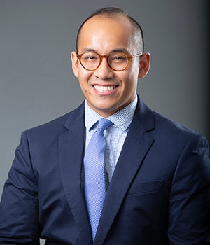 A balding man with a ring of black hair wears a blue business suit and rust-colored glasses while smiling.