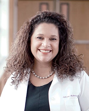 A woman with curly brown hair wearing a beaded necklace and a white lab coat smiles at the camera.