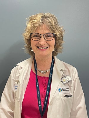 Older nurse with short graying blond hair wearing black-framed glasses and a fuchsia shirt underneath a lab coat. 