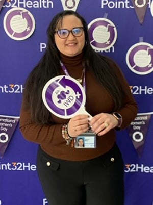 A woman with long brown hair, glasses, and a brown turtleneck stands in front of a purple wall at a Point32health event holding a sign that says "IWD." 