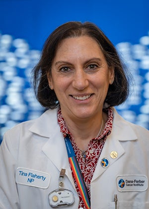 Smiling nurse with shoulder length brown bob wearing a lab coat over a swirly red patterned blouse and a rainbow lanyard around her neck. 