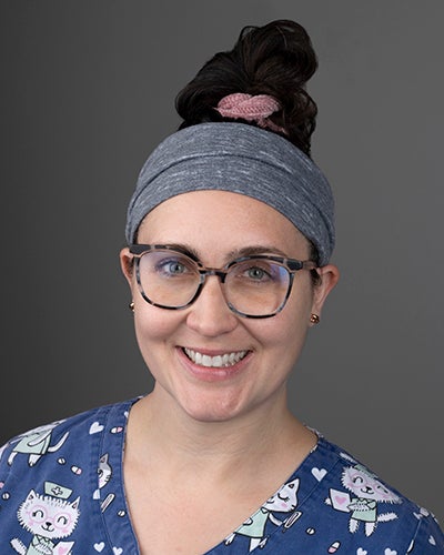 Quirky nurse with tortoise shell glasses wearing dark hair up in a topknot cinched with a pink scrunchy and a thick grey sweatband. 