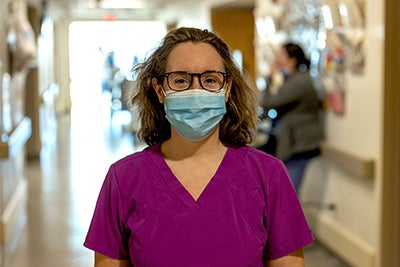 Nurse with wavy hair pulled back with headband wears mauve scrubs and stands in hospital hallway