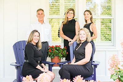Two women in black dresses sitting in blue adirondack chairs in front of an older man and two more women in black dresses standing up. 