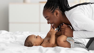 A happy Black mom leans over her baby and holds his feet, as he touches her face curiously. 