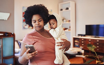 A Black mom in a pink T-shirt holds her baby in one arm and looks at her phone with her other hand.
