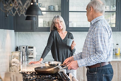 Senior man cooking breakfast in a frying pan and talking to his wife, who is holding a cup of coffee and smiling. 