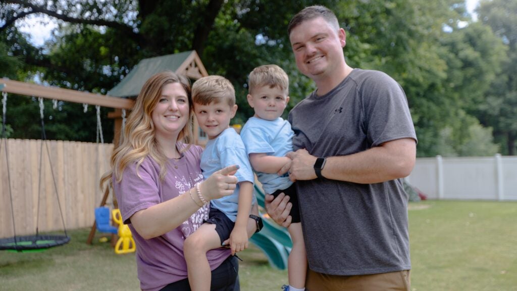 Kelsey, her husband, and their two sons pose for a family photo in their backyard. In the background they have a swingset.