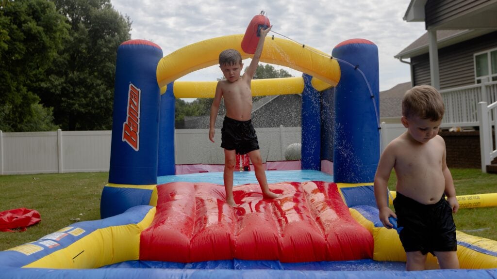 Colt and his younger brother play on an inflatable water toy in their backyard. Colt points towards the sky with an inflatable red water toy. 