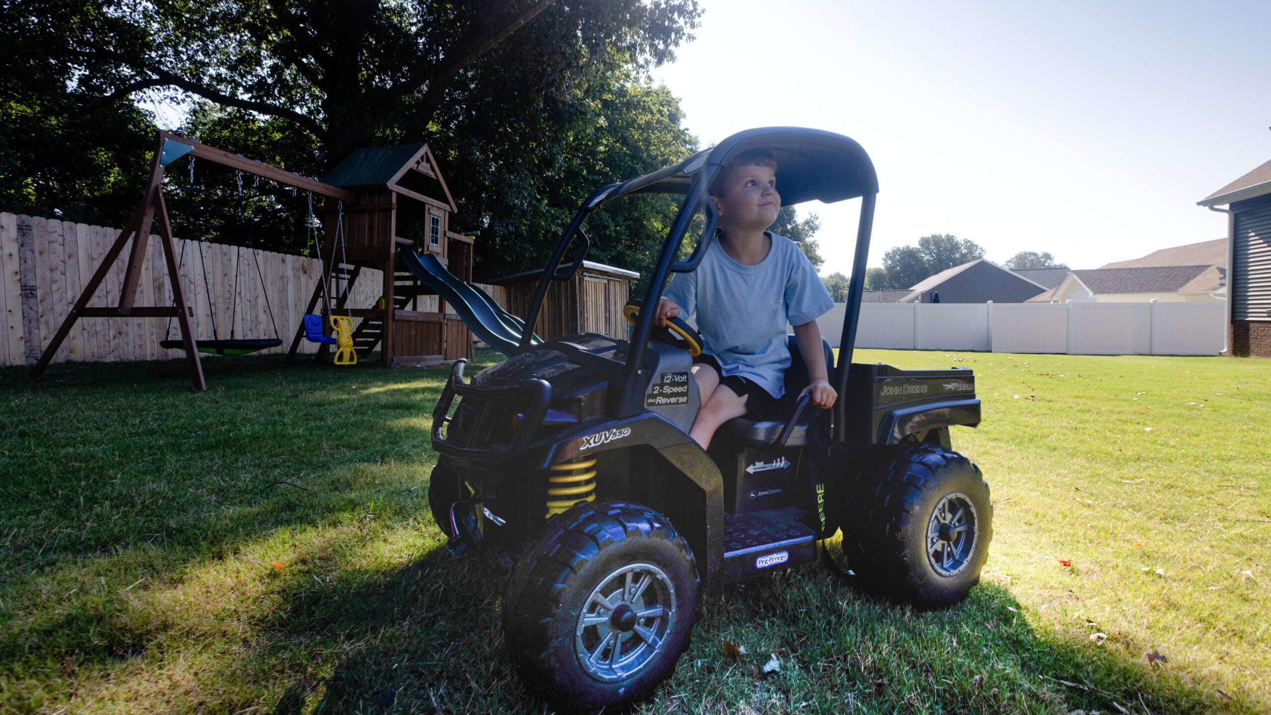 Colt looks up at the sky whilst riding a child-sized toy truck around his backyard. A jungle gym, and a verdant green lawn lie in the background.