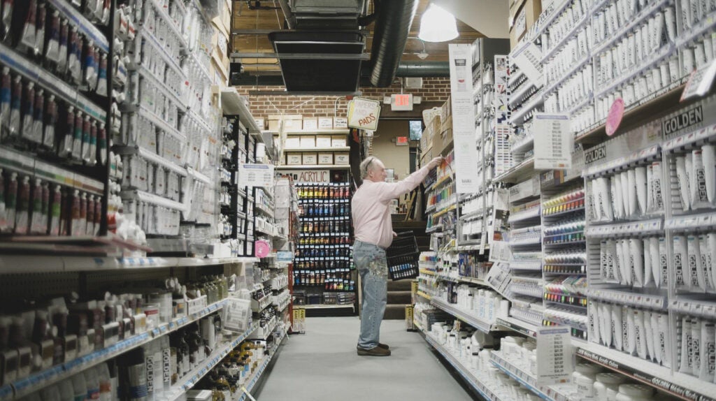 Older man reaching for a tube of paint in the paint aisle of an art supply store. 