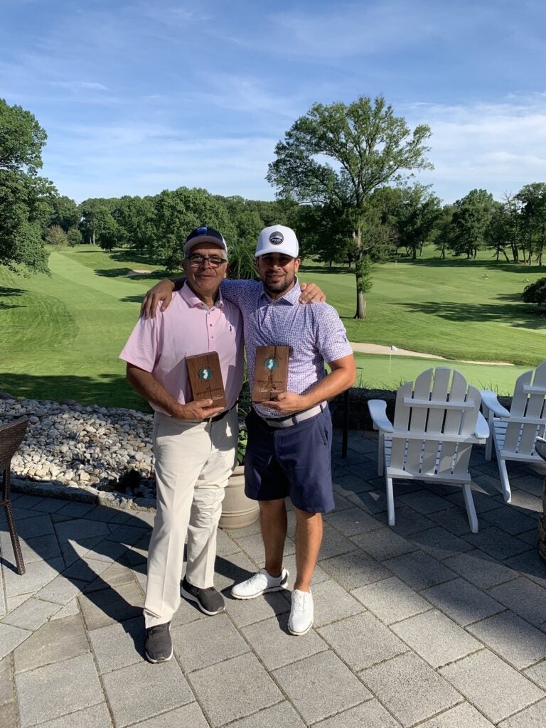 Recent photo of Tony Gallardo and his adult son posing on a golf course while holding plaques and smiling with their arms around each other. 