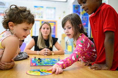 Three children playing a board game while blond teacher watches.