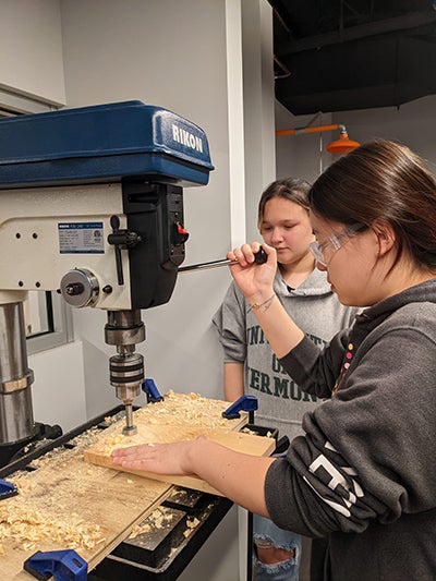 Two girls working with a large machine in wood shop. 