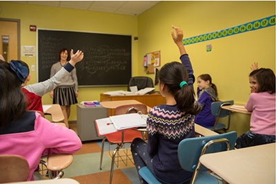 Students in yellow classroom learning and raising their hands to answer their teacher's question. 