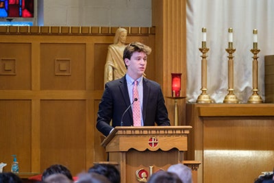 Blond young man giving a speech at the podium in his school's chapel. 