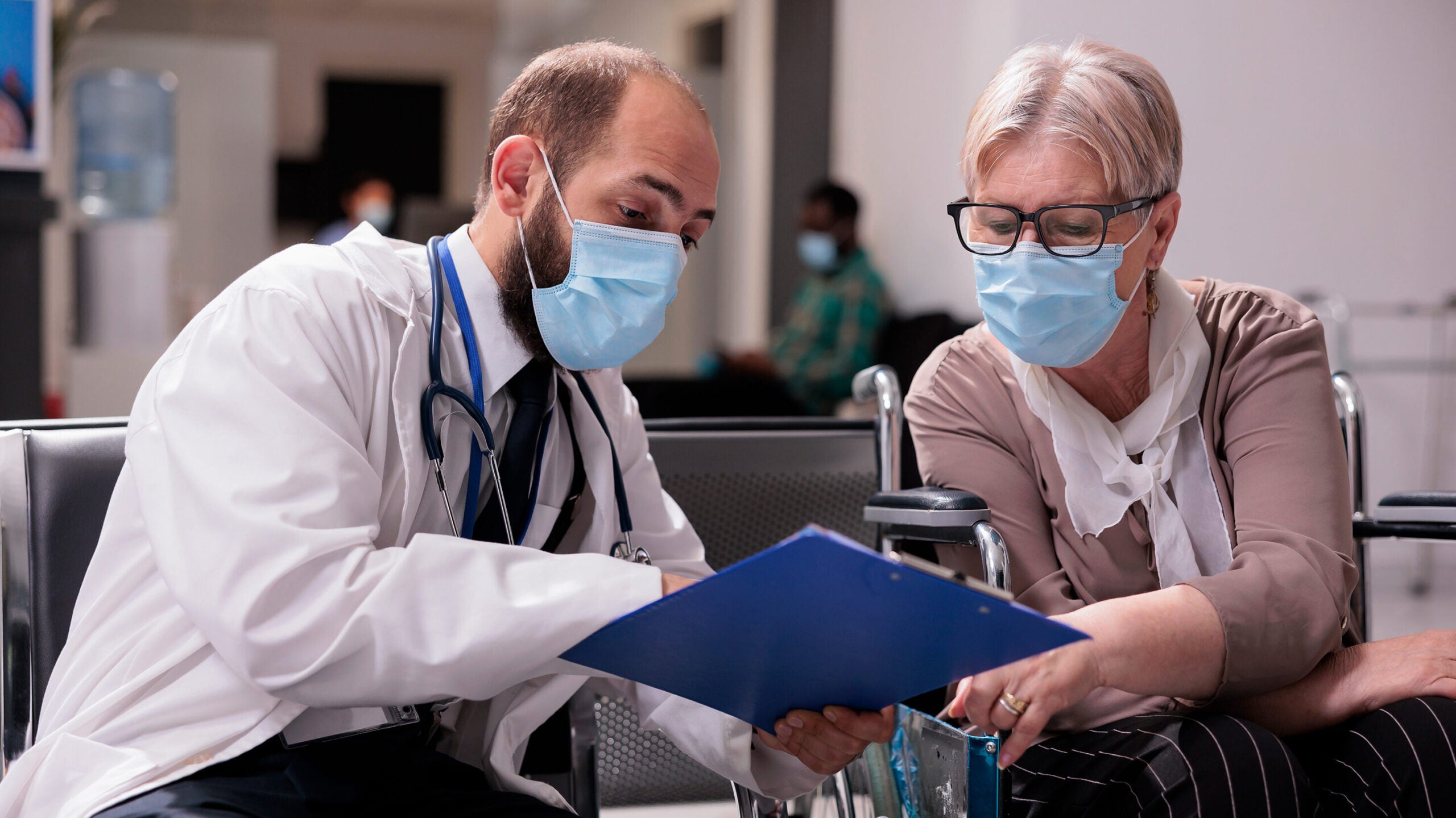 A male doctor sits down with an older female patient to discuss her medical history. They are both wearing masks and looking down at the doctor's blue clipboard. 