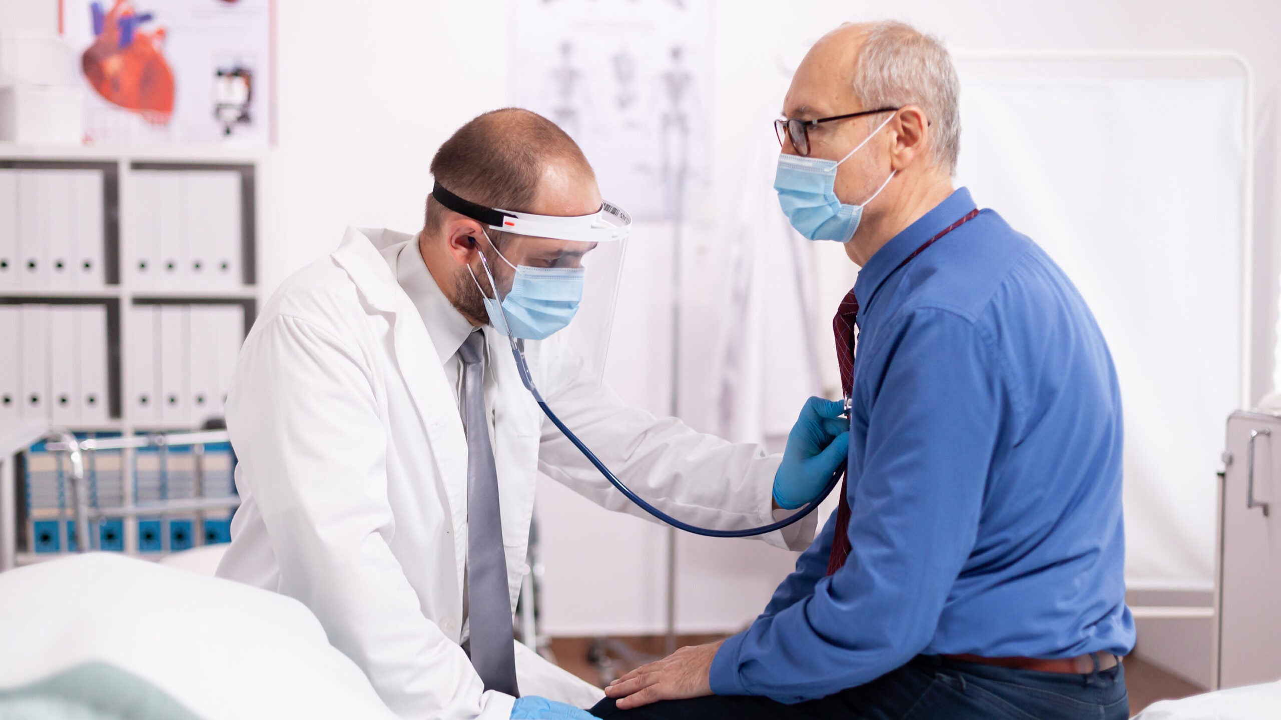 A male doctor checks the heartbeat of an older male patient with a stethoscope. They are both sitting down. 