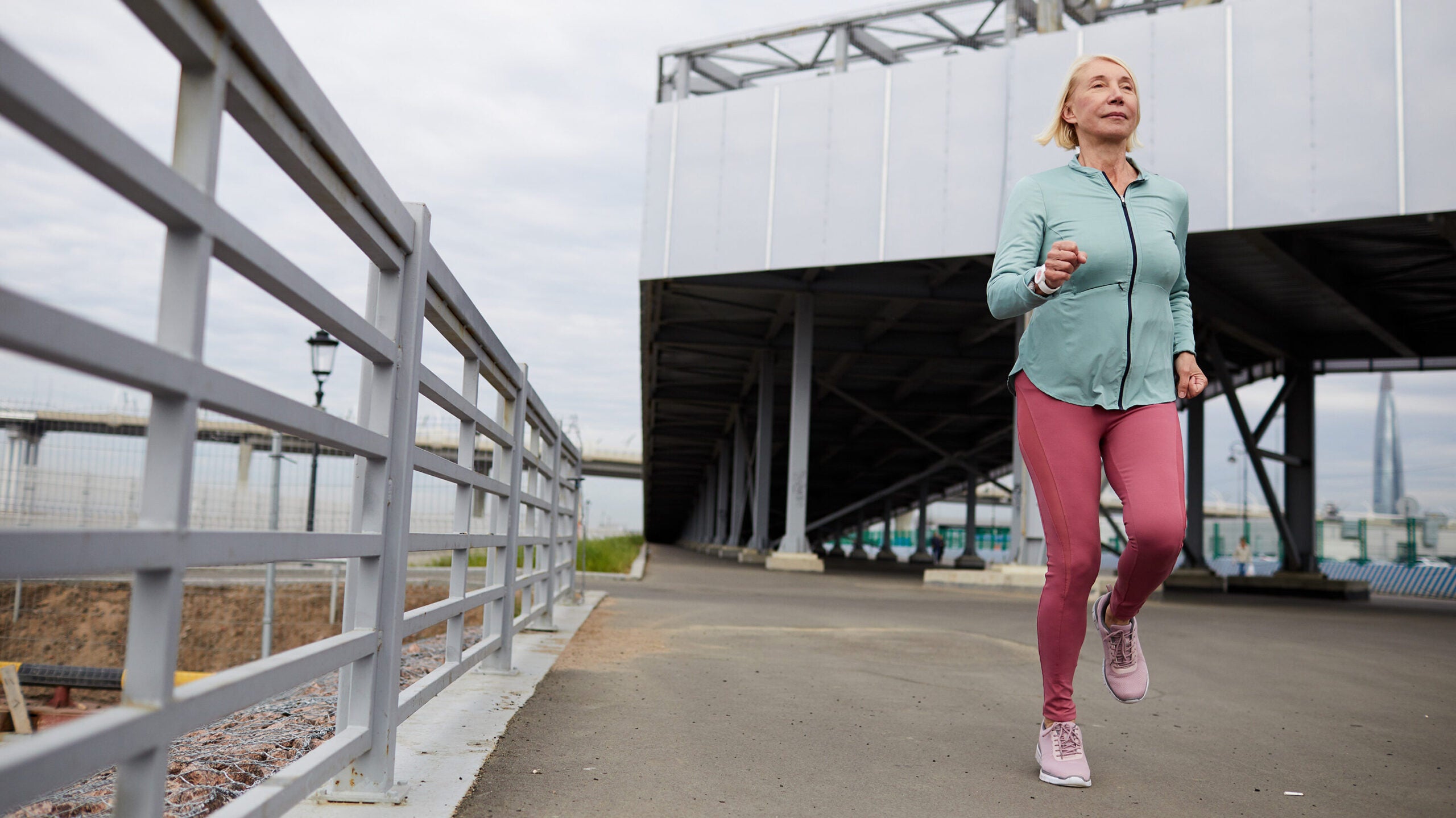 An older woman wearing leggings and a sweatshirt jogs on a grey bridge.