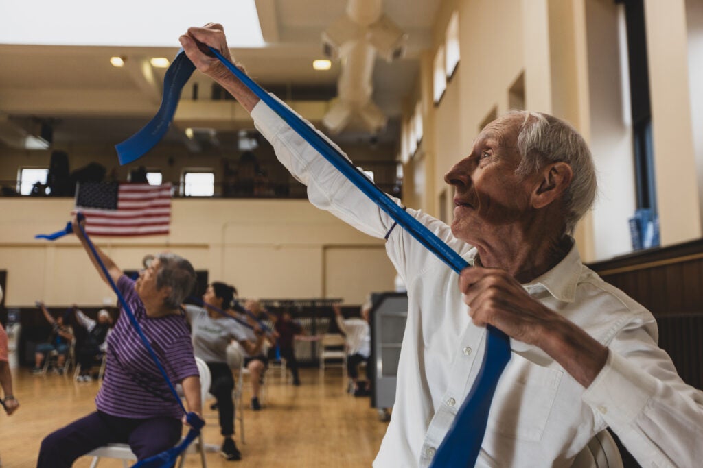 An older adult man stretches a workout band to the ceiling while several other older adults do the same motion in the background during a senior community exercise class. 