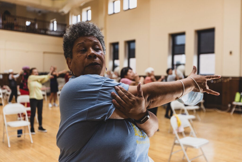 An older adult woman stretches her right arm across her body while several other older adults do the same motion in the background during a senior community exercise class.