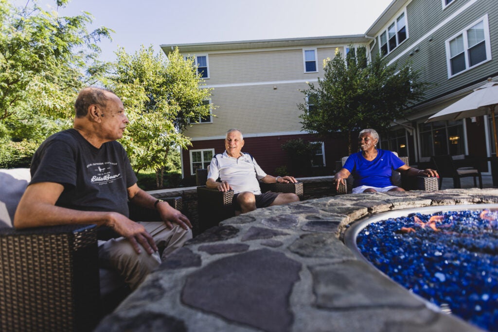 Three older adult men talk and smile to each other as they sit outside at a senior living community center on a sunny day. 