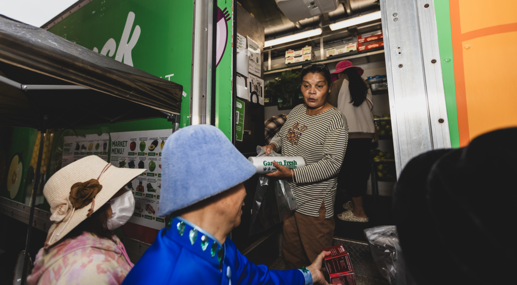 A woman stands in the Fresh Truck and holds a roll of plastic produce bags. In front of her, two people stand, wearing hats, talking to her. 