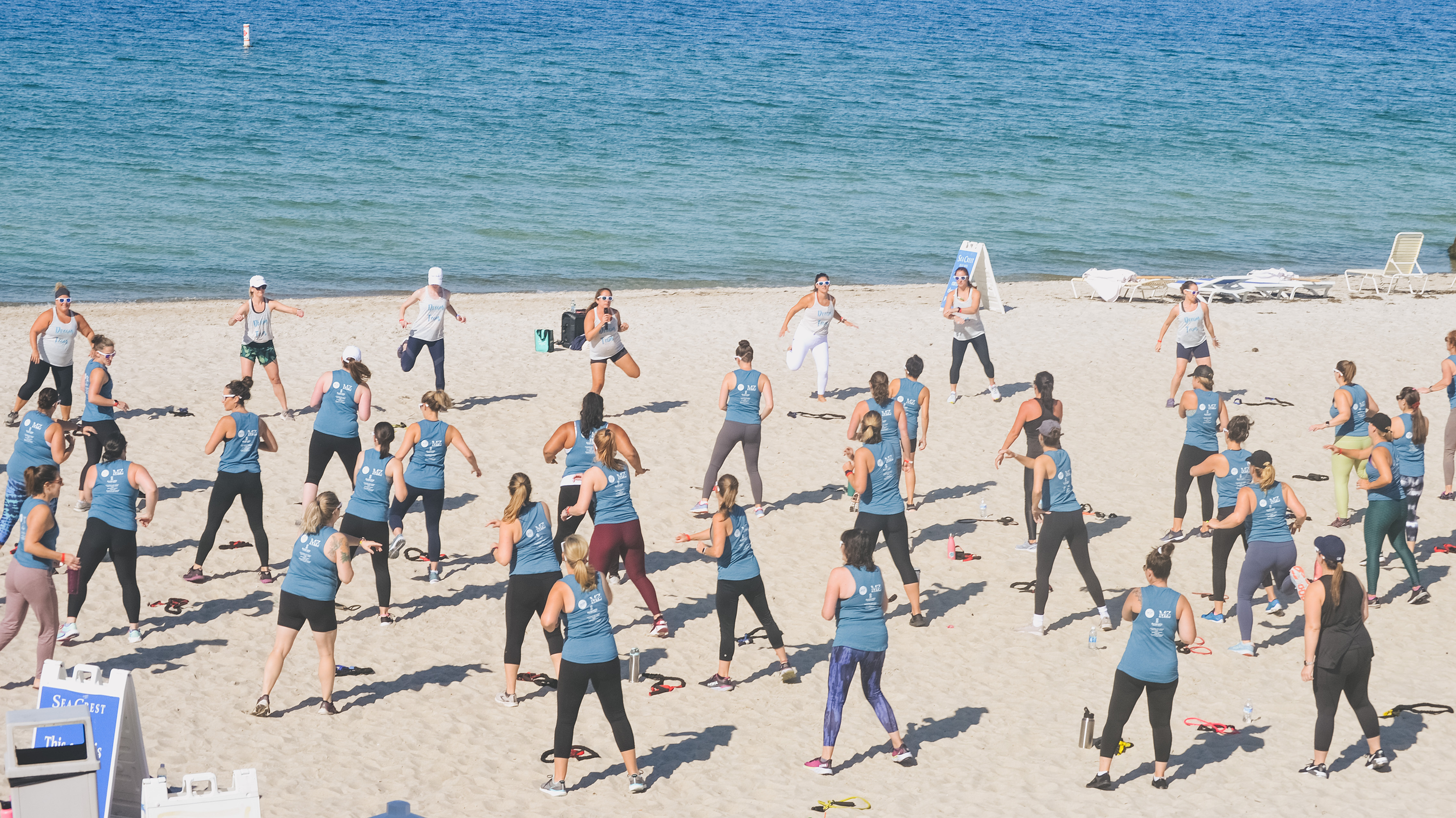 The Mama Beasts group of about 30-40 women in matching blue shirts participate in a group fitness class on the beach on a bright, sunny day. 