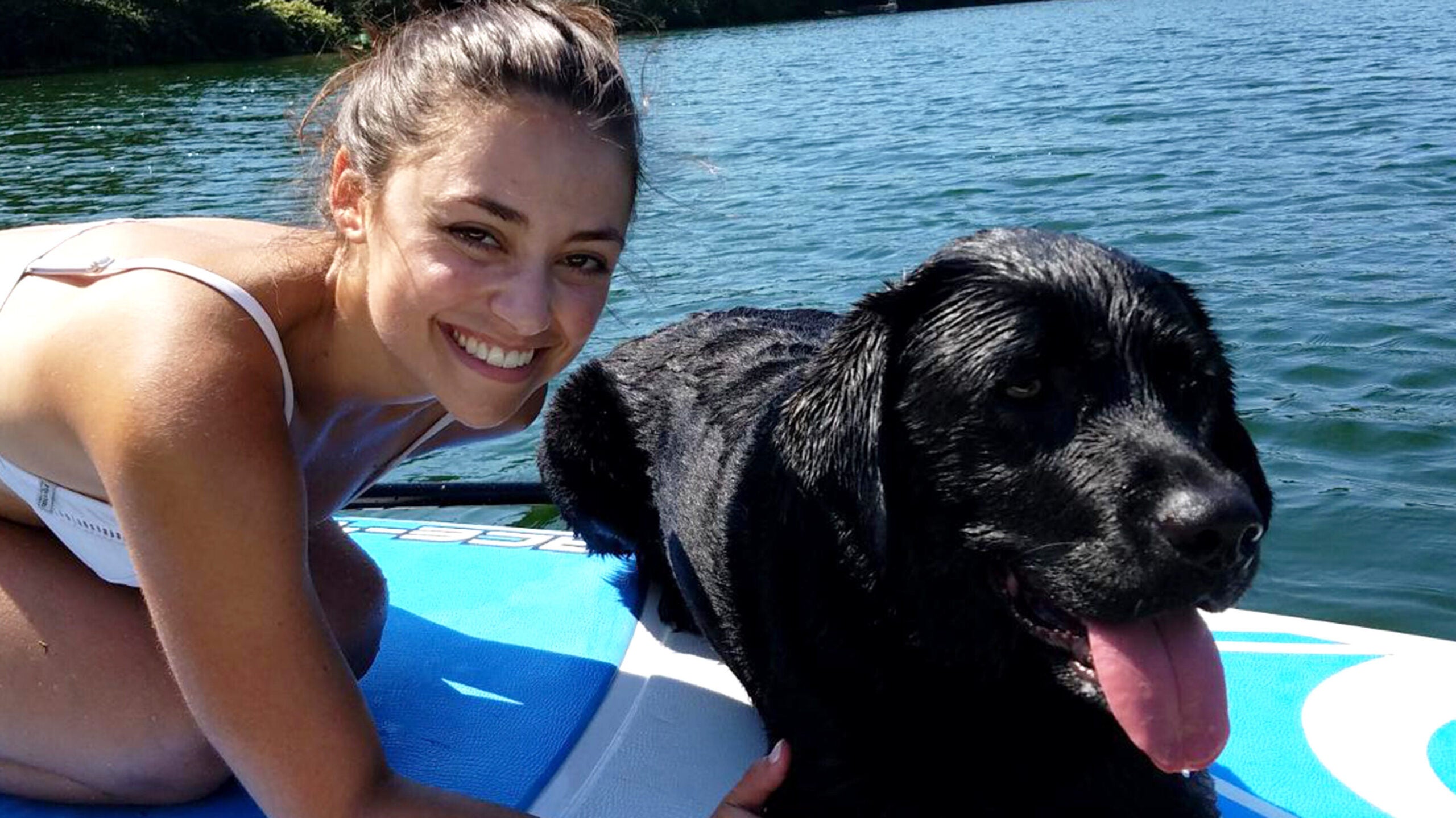 Liz and her black labrador retriever, Riley, pose for a photo on a paddleboard on a lake. 