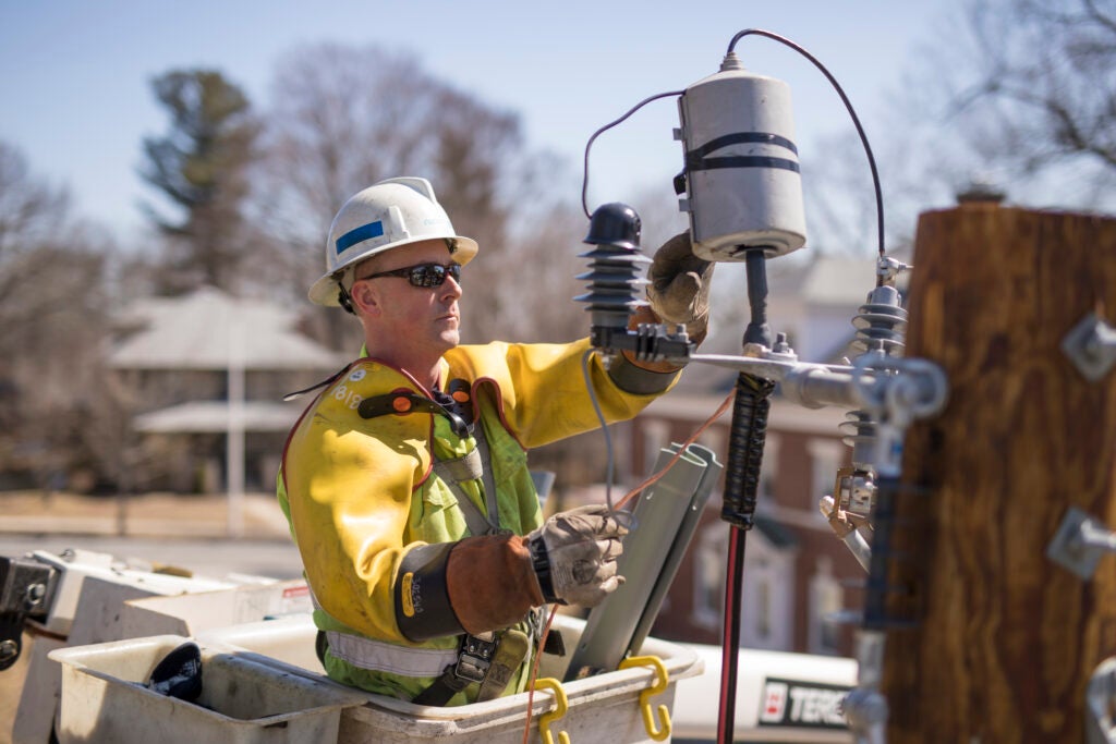 A National Grid employee wearing a yellow work jacket, a hard hat, and sunglasses does electrical work in a residential neighborhood