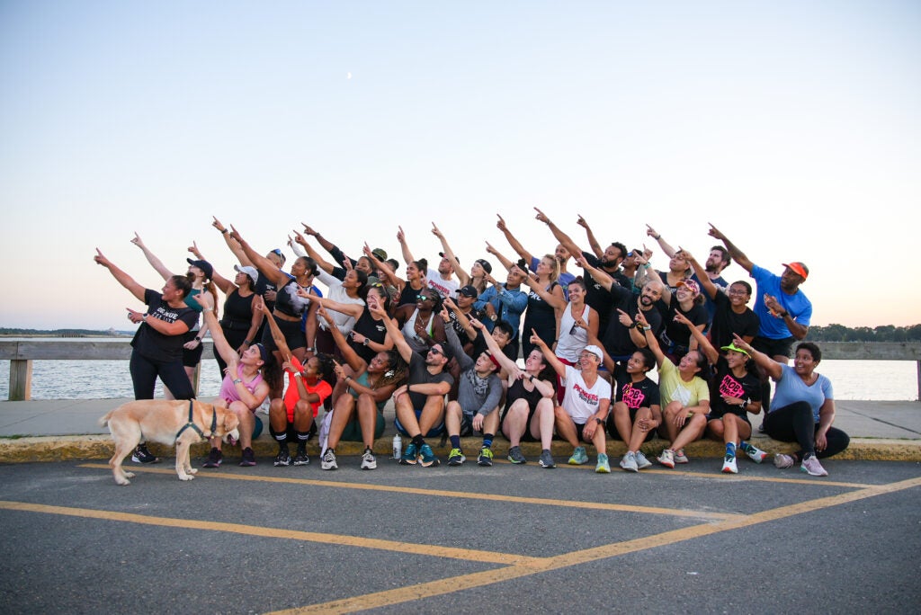 Members of the Pioneers Run Crew pose in front of a reservoir together, stretching each of their hands up in unison to point at the sky. 