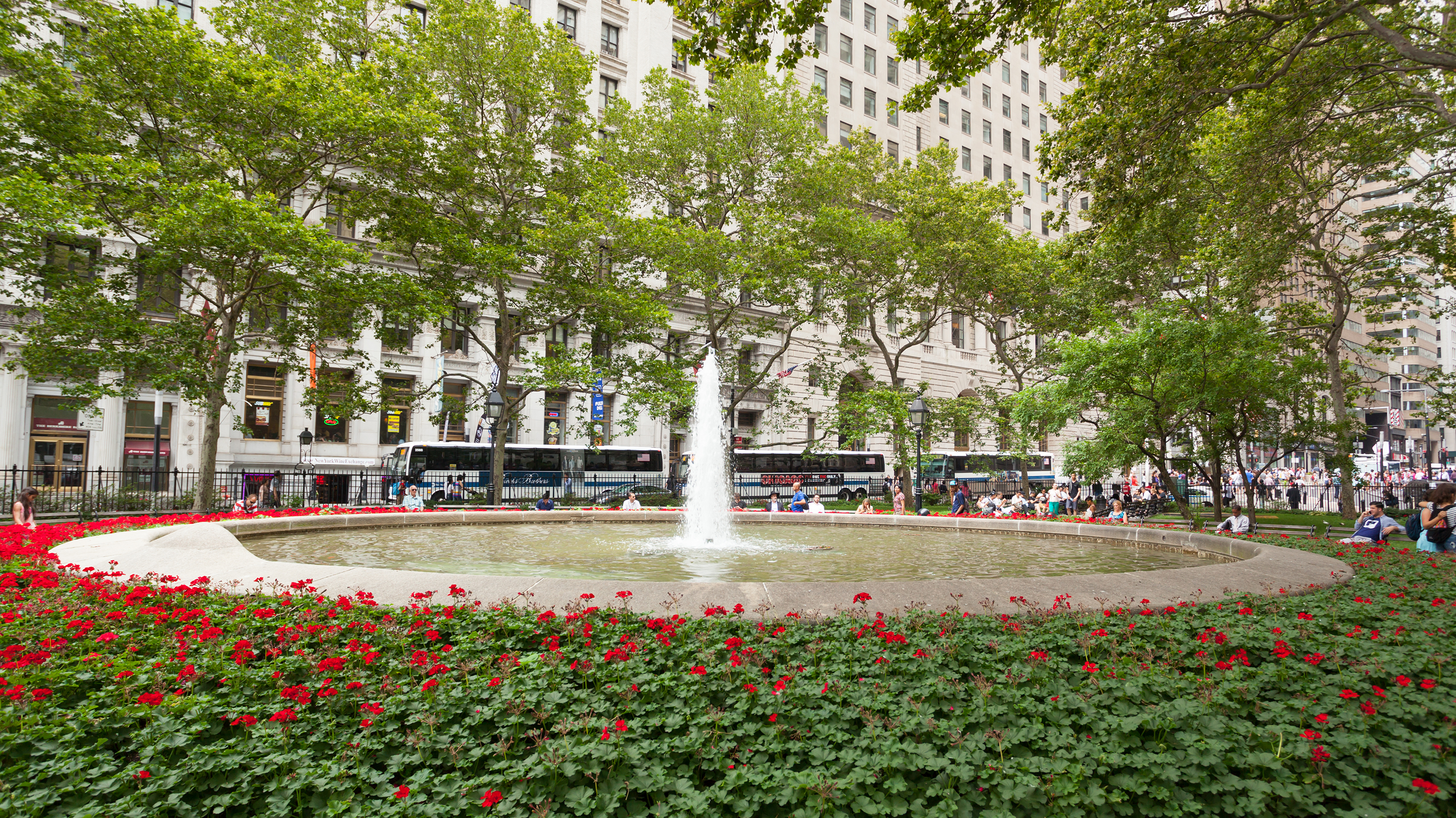 In the middle of a city, there is a green space filled with trees, red flowers, and in the middle, a flowing fountain. 