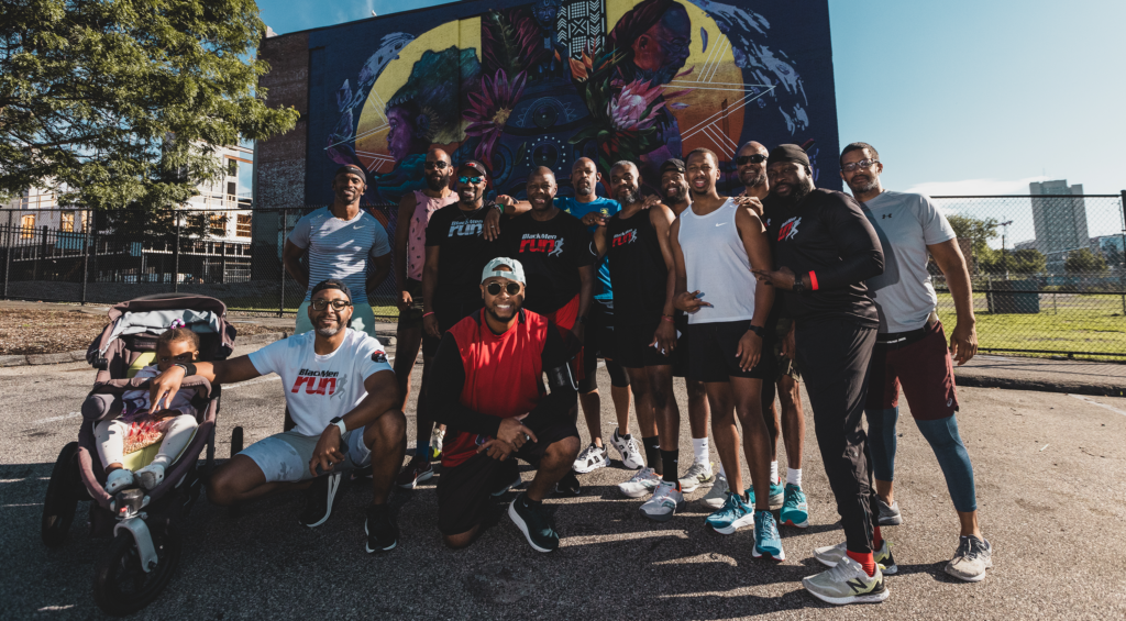 Thirteen members of Black Men Run's Boston chapter pose for a photo on a blacktop with a colorful mural in the background. 