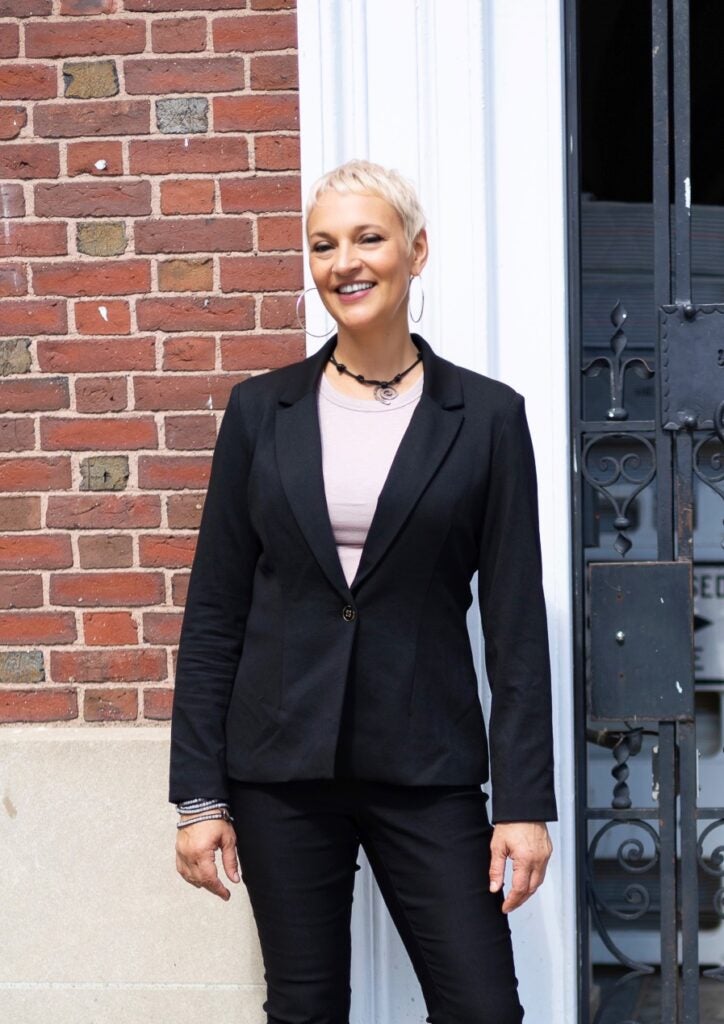 A woman with short, light hair poses wearing a black blazer and slacks in front of a brick building. 