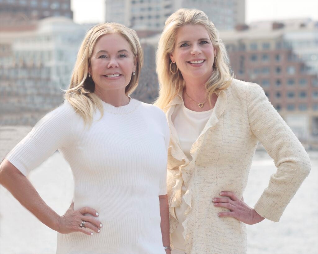 Two women with blonde hair wearing professional attire pose together in front of a cityscape.