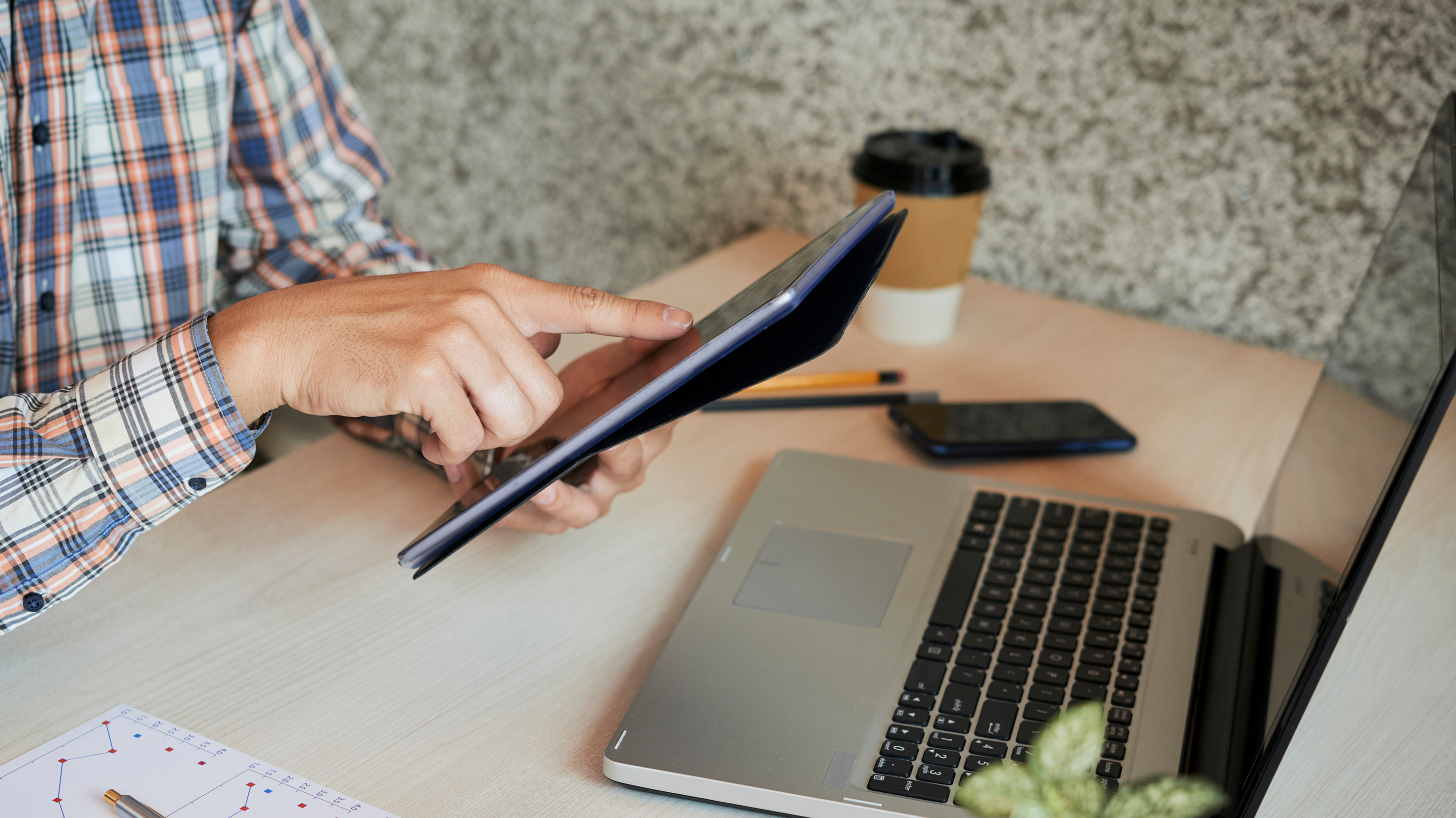 A man sits at a table with a coffee, his laptop, a tablet, some pencils, and his phone. He is scrolling on the tablet with his finger, pointing to something on the screen.