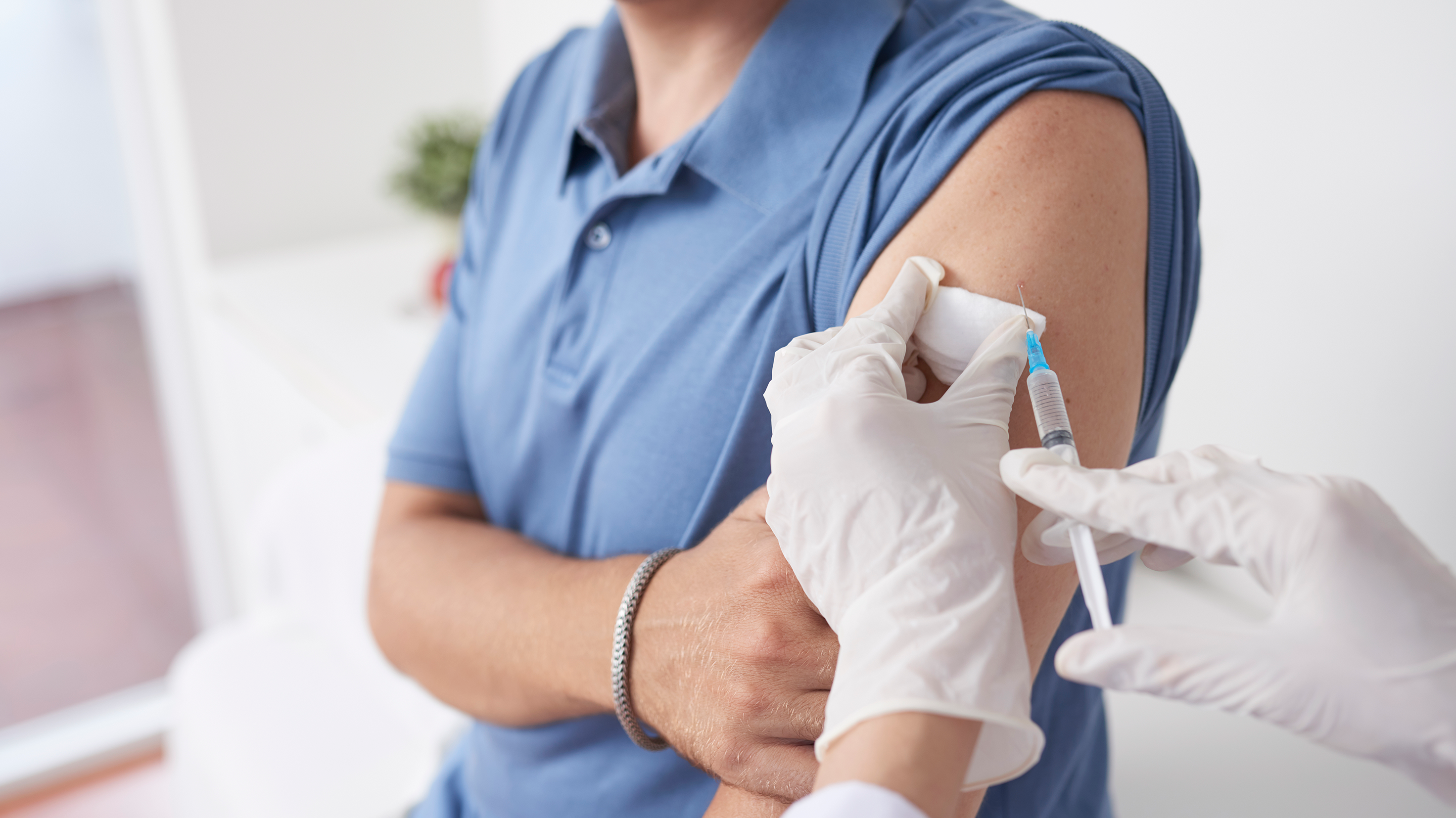 A man in a blue shirt sits while getting an injection by a doctor, wearing white latex gloves.